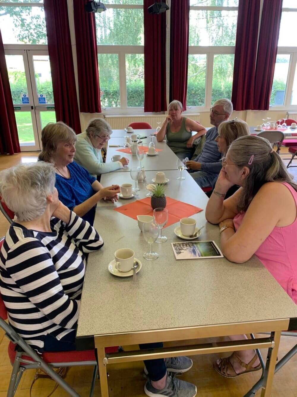 A group of people sitting around a table with cups and saucers, engaged in conversation, in a brightly lit room. - Home Instead