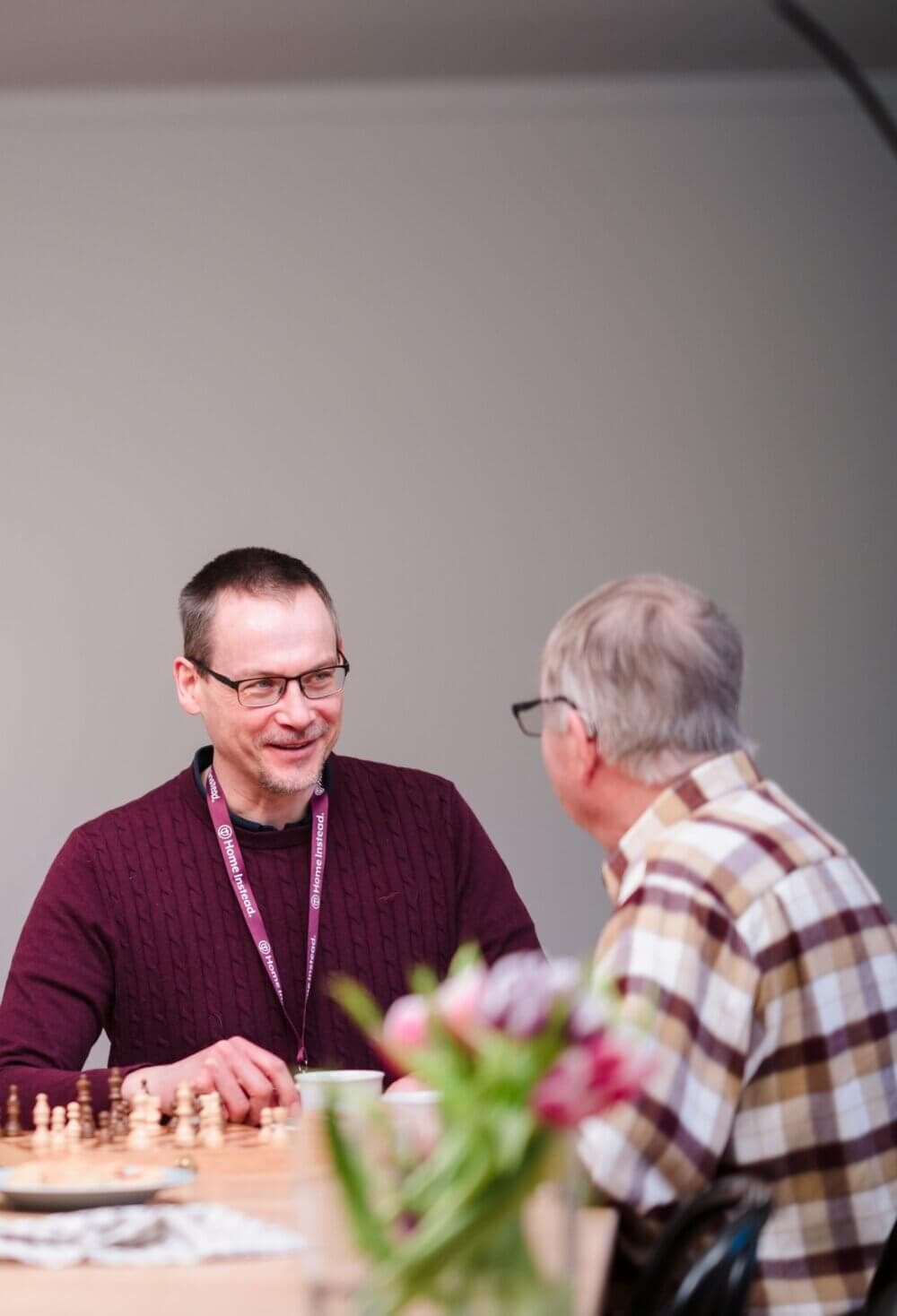 Two men having a conversation over a chess game; one is smiling, wearing glasses and a maroon sweater. - Home Instead Bournemouth & Christchurch