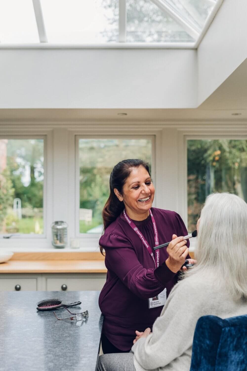 A caregiver wearing a purple top and name tag applies makeup to an elderly woman in a well-lit room with large windows. - Home Instead