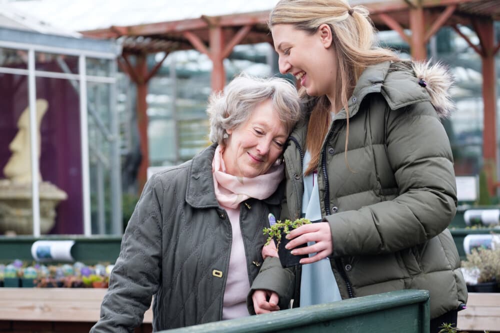 Two women in jackets, one elderly and one younger, smile and bond while holding a small plant in a garden center. - Home Instead