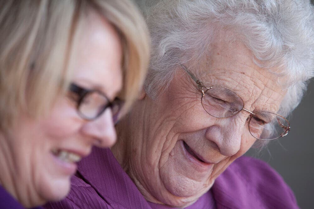 Two elderly women, one with glasses, smiling and looking down while standing close together. - Home Instead