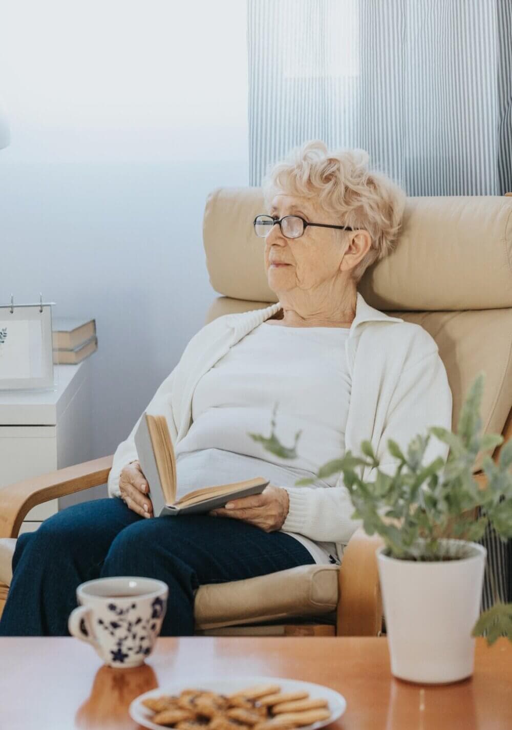 Elderly woman with glasses reading a book in an armchair, next to a table with mug, plate of cookies, and a plant. - Home Instead