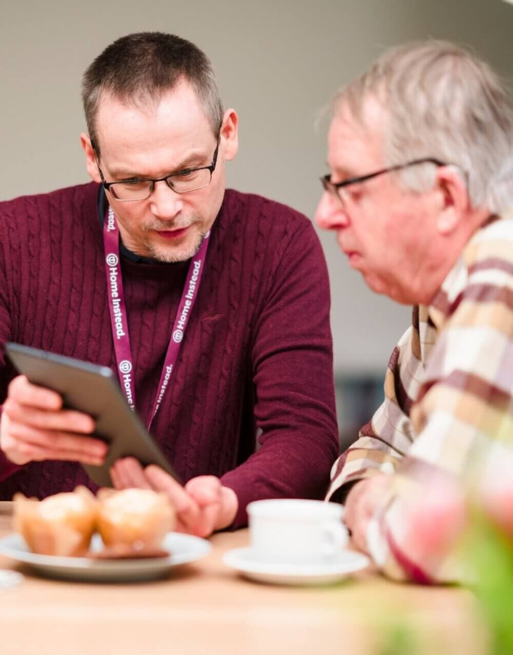Two men at a table, one showing the other something on a tablet. Muffins and a cup of coffee are on the table. - Home Instead