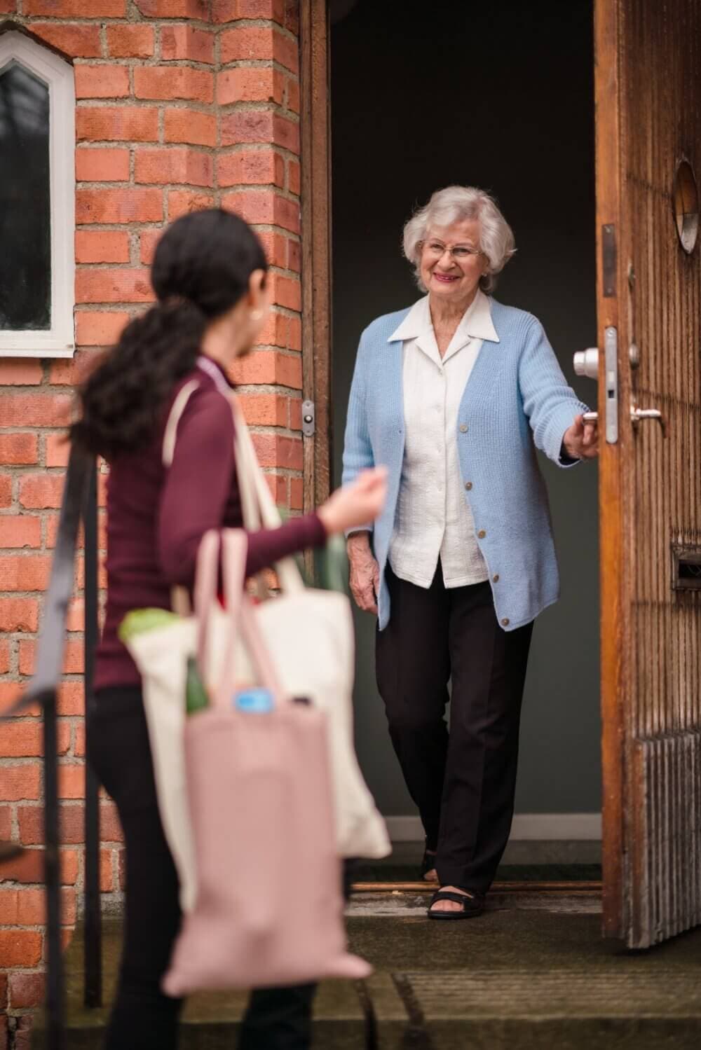 Older woman holding door open for person with bags outside a brick building. - Home Instead
