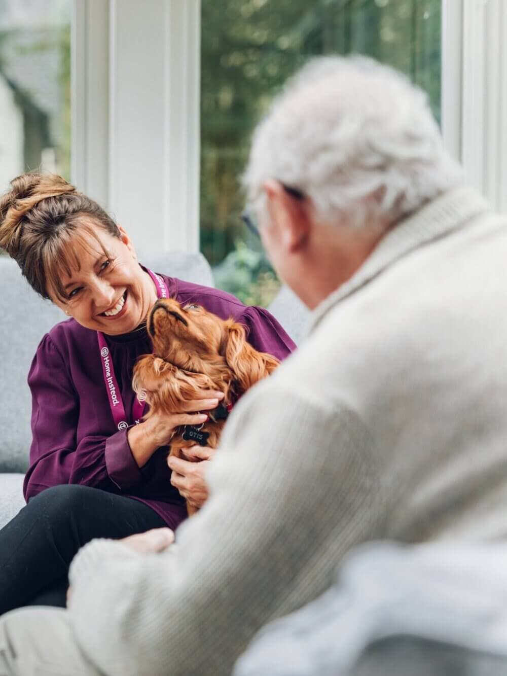 A woman with a dog and an elderly man are smiling at each other while sitting on a couch. - Home Instead
