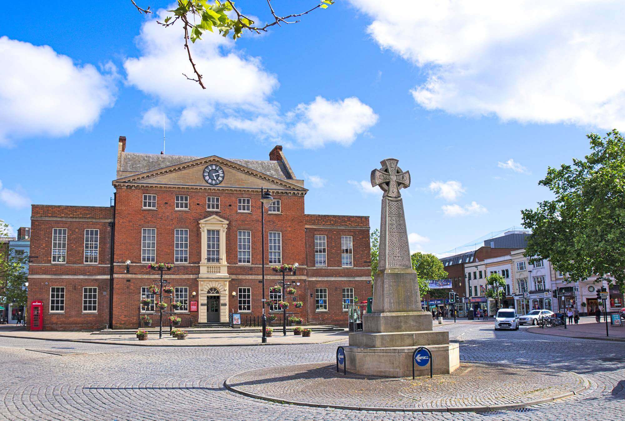 A historic brick building with a clock tower and a stone cross monument in a sunny town square. - Home Instead