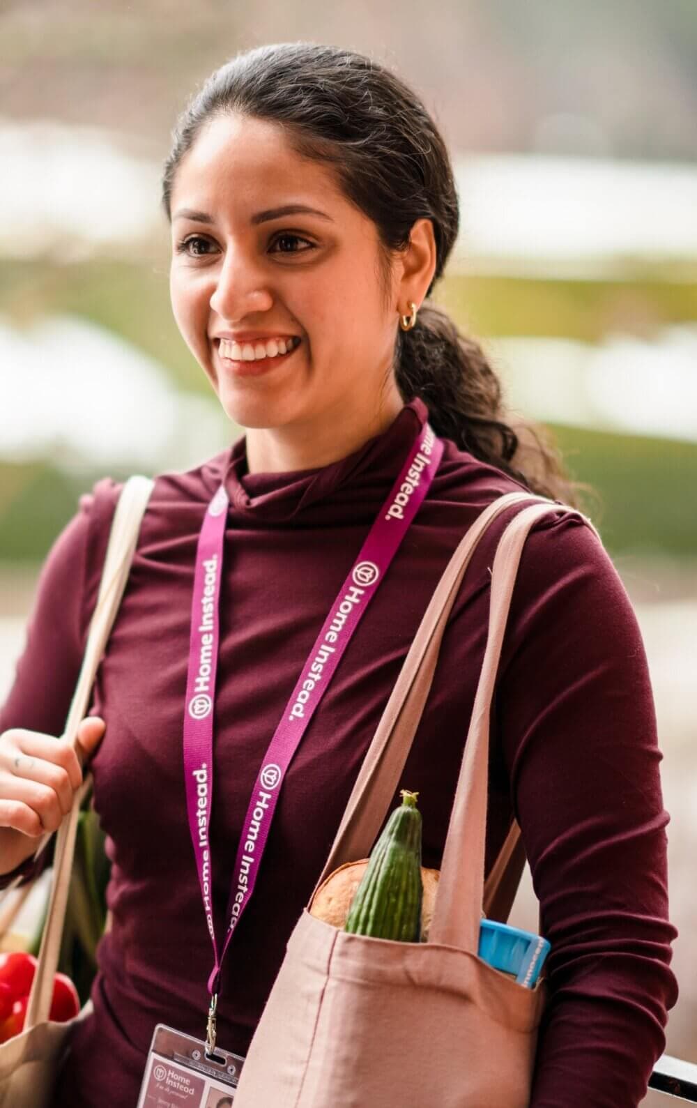 A smiling woman carrying reusable grocery bags, wearing a lanyard with a badge. - Home Instead Poole
