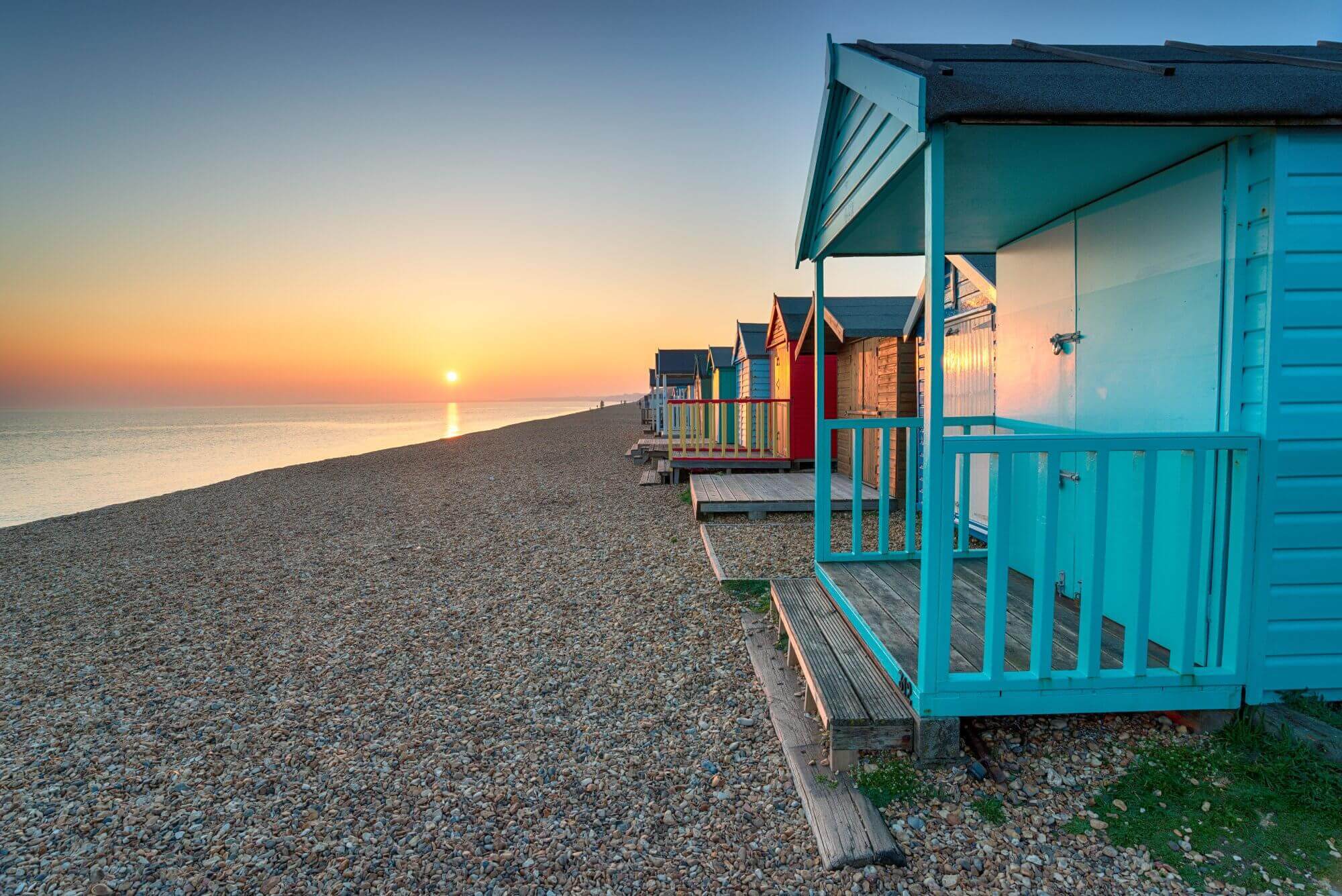 Colorful beach huts lined up on a pebble beach at sunset, with the sun setting over the calm sea in the background. - Home Instead