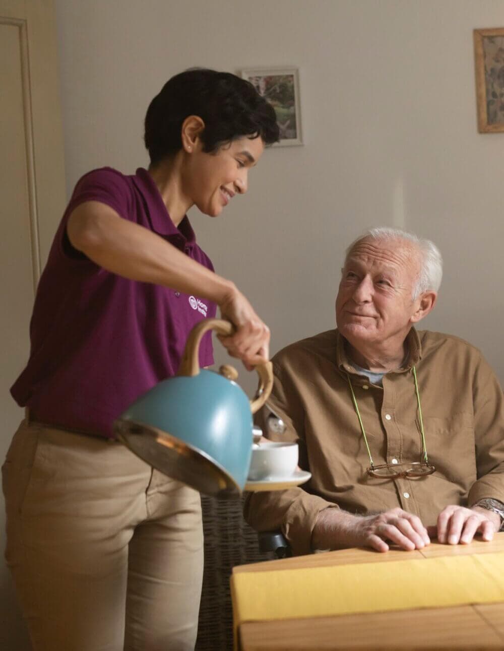 A caregiver in a purple shirt pours tea for an elderly man at a table, both smiling at each other. - Home Instead