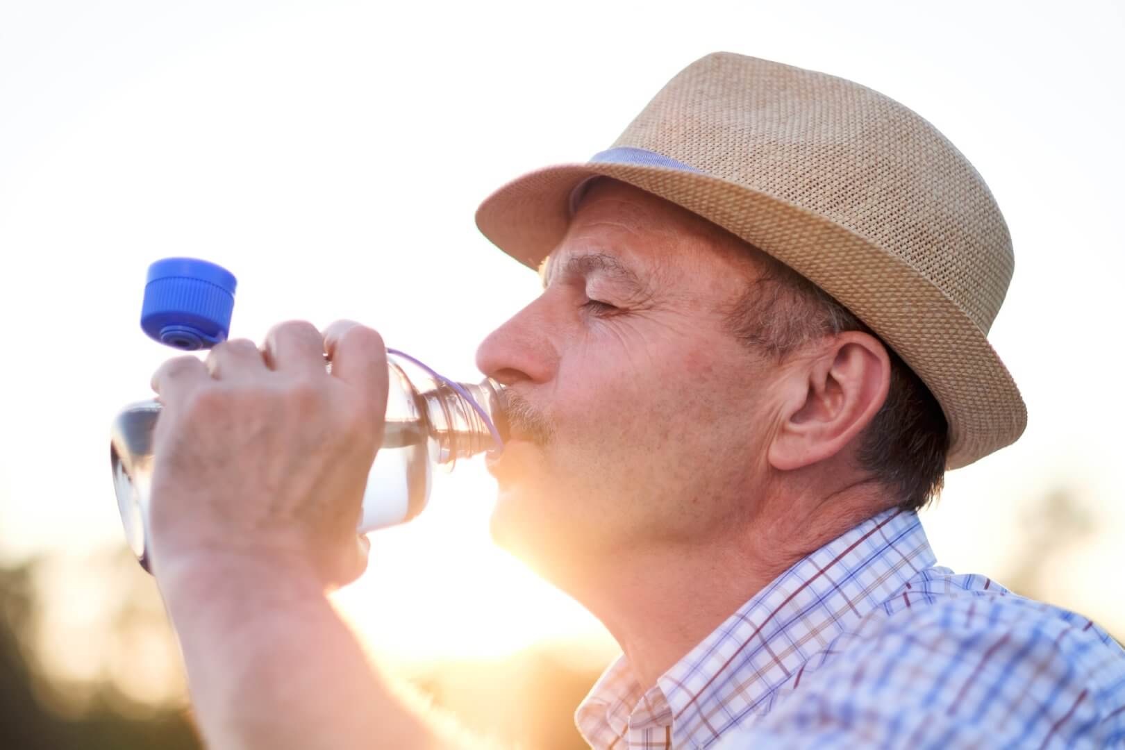 A man wearing a hat drinks water from a bottle outdoors, with the sun setting in the background. - Home Instead