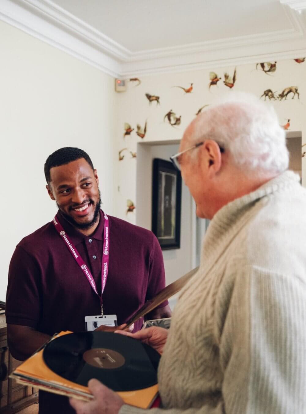 A young man in a maroon shirt helps an older man wearing a beige sweater hold a vinyl record. - Home Instead