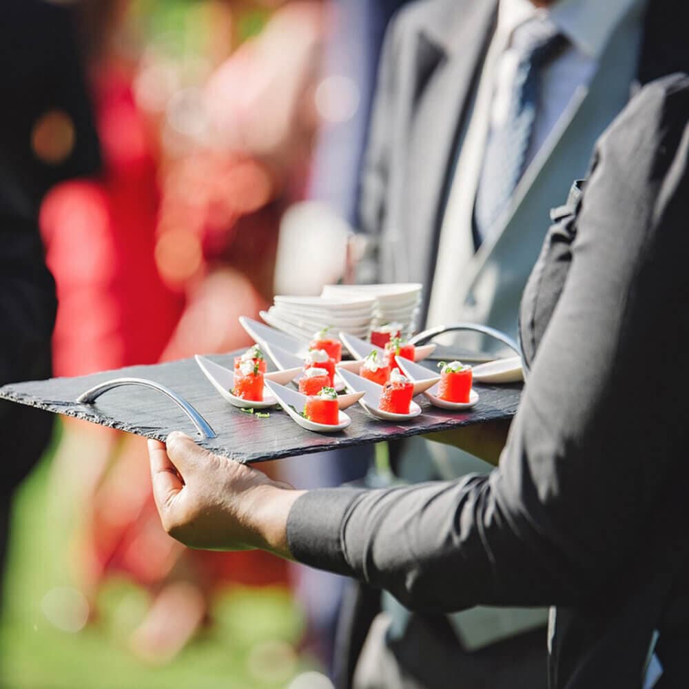 A server holds a tray with small appetizers on white spoons at an outdoor event. People in suits are in the background. - Home Instead