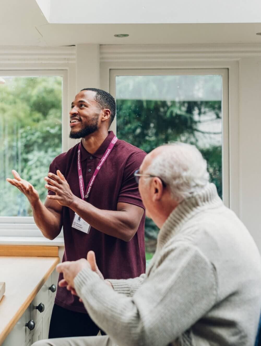 Young man in a maroon shirt converses with an older man in a gray sweater in a brightly lit room. - Home Instead