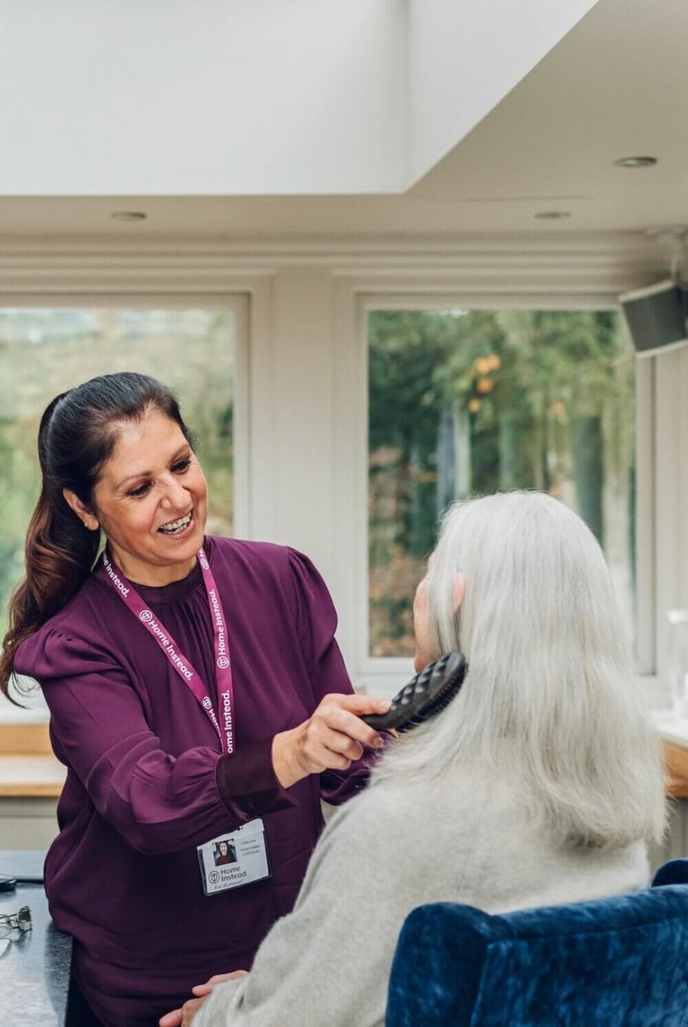 A woman brushes an elderly woman's hair in a sunlit room, both are smiling. The younger woman wears a name tag. - Home Instead