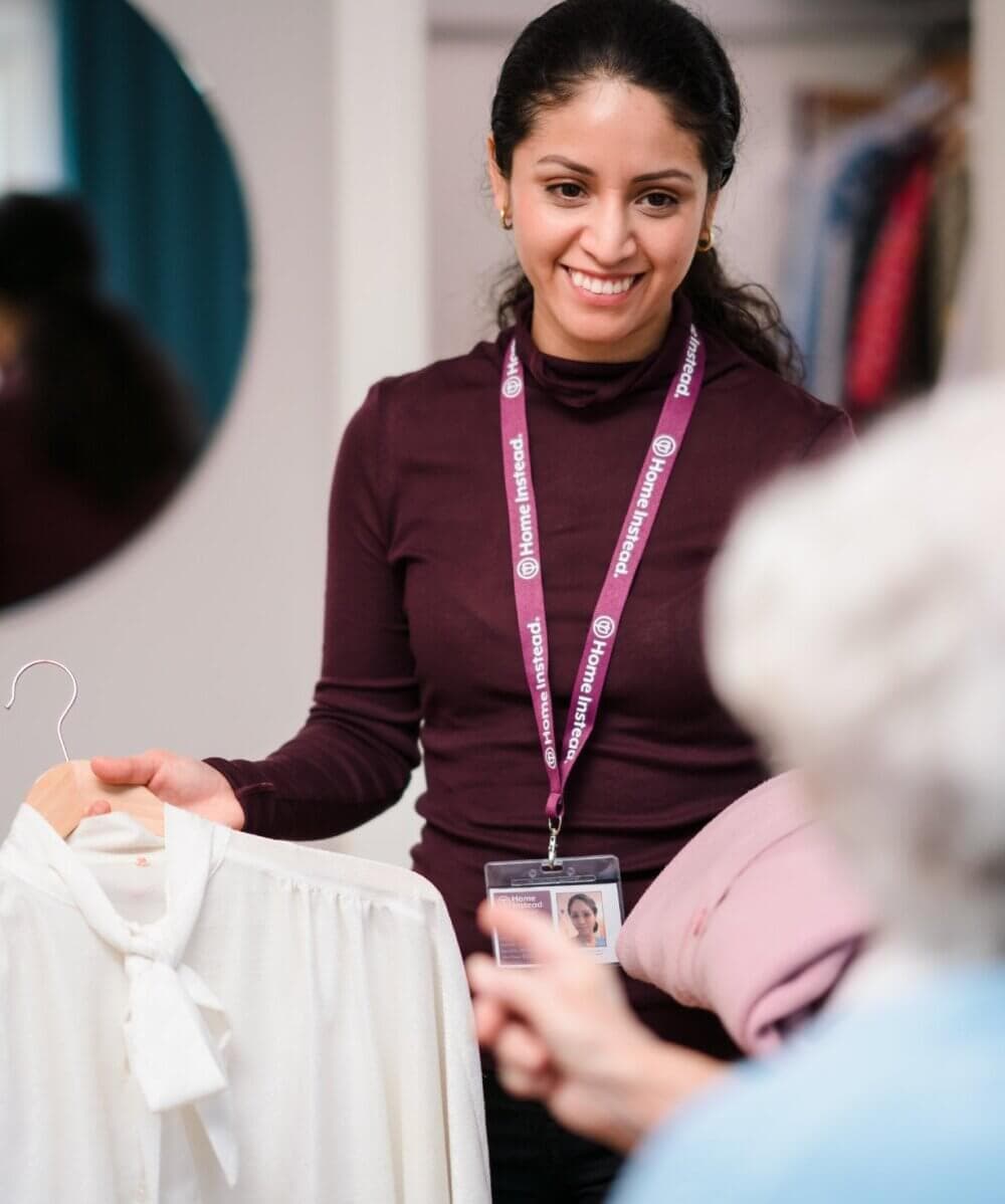 A woman, smiling and holding a white blouse on a hanger, shows it to someone out of frame while wearing an ID badge. - Home Instead