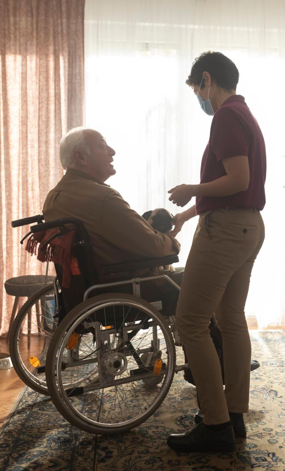 A person in a wheelchair holds a small dog while a caregiver stands beside them, both wearing masks in a sunlit room. - Home Instead