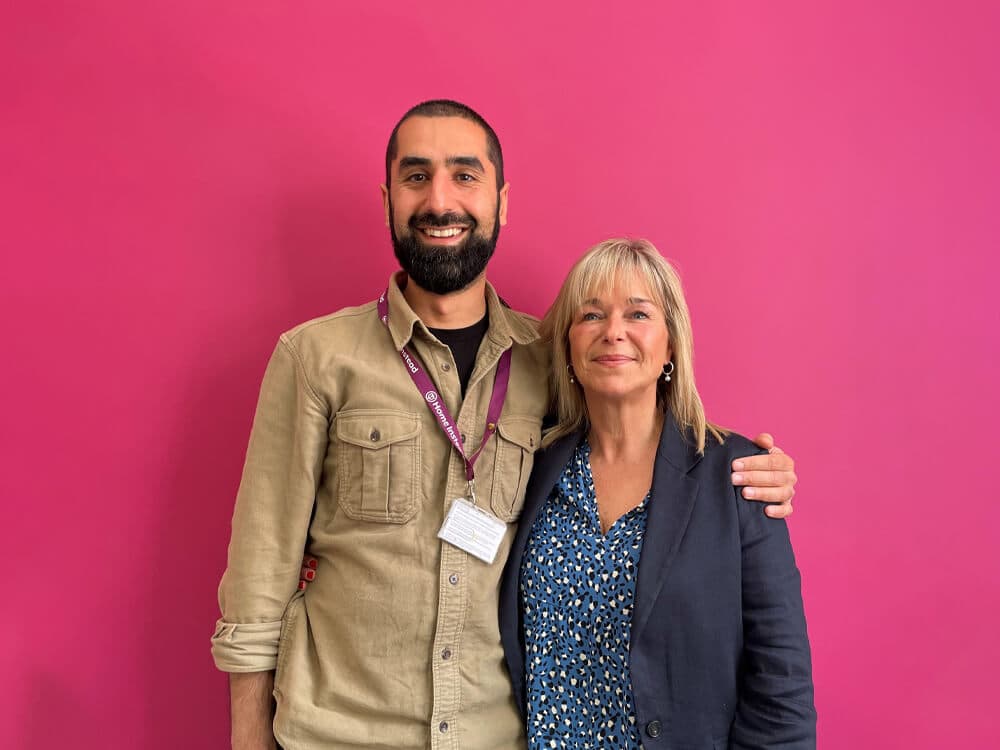 Two people smiling in front of a bright pink background. One is wearing a beige shirt, the other a blue patterned blouse. - Home Instead