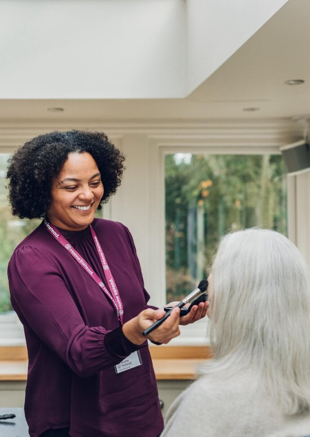 A caregiver smiling while applying makeup to an elderly person with gray hair in a bright room. - Home Instead