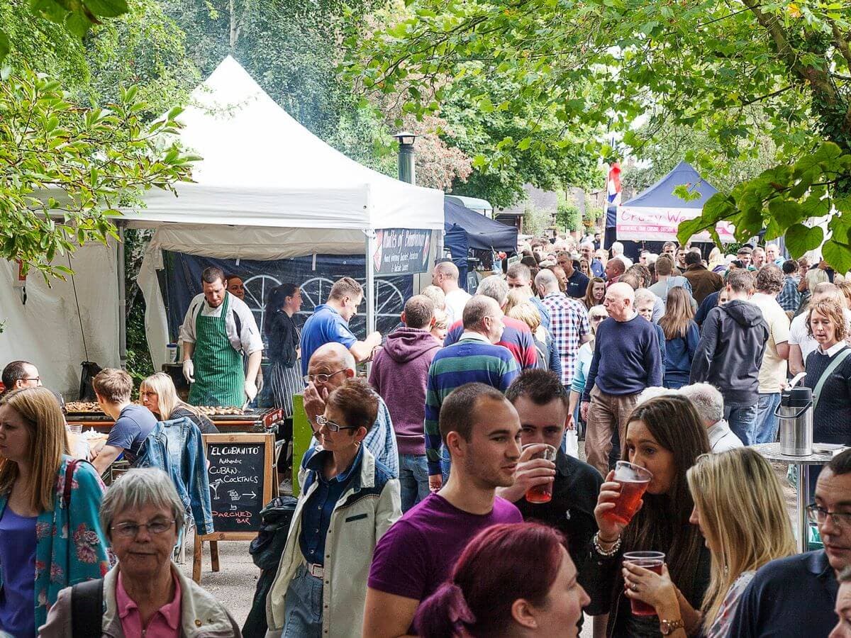 A crowd of people enjoying an outdoor market with food stalls and greenery in the background. - Home Instead