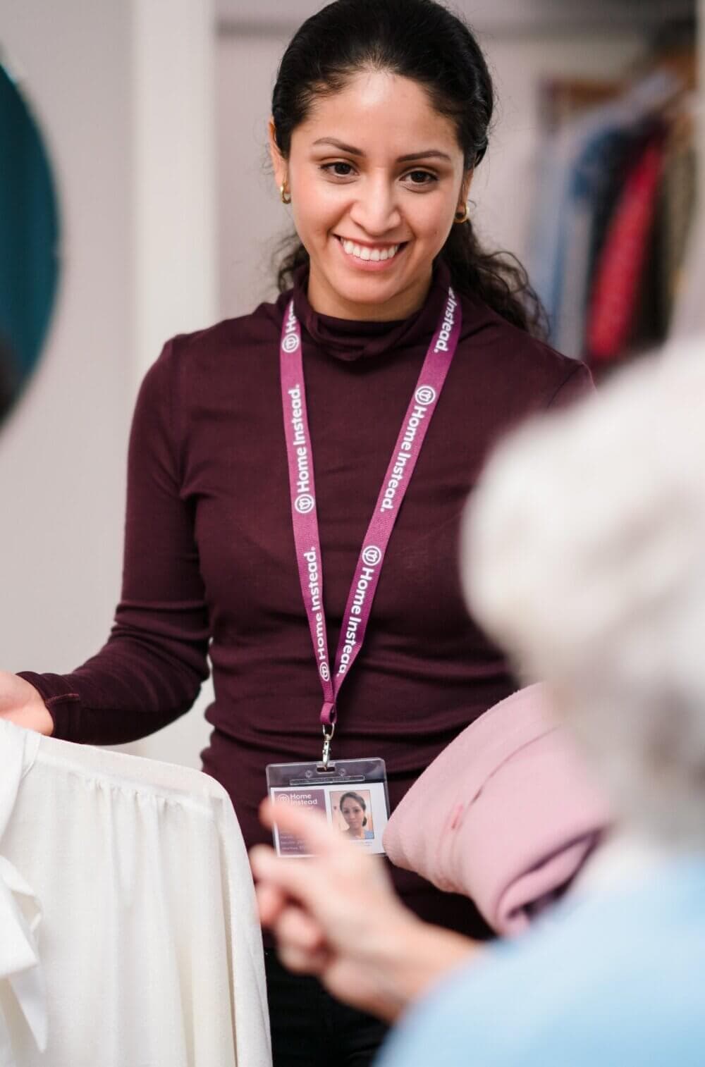 A smiling woman with a lanyard assists an older person with clothes shopping in a store. - Home Instead