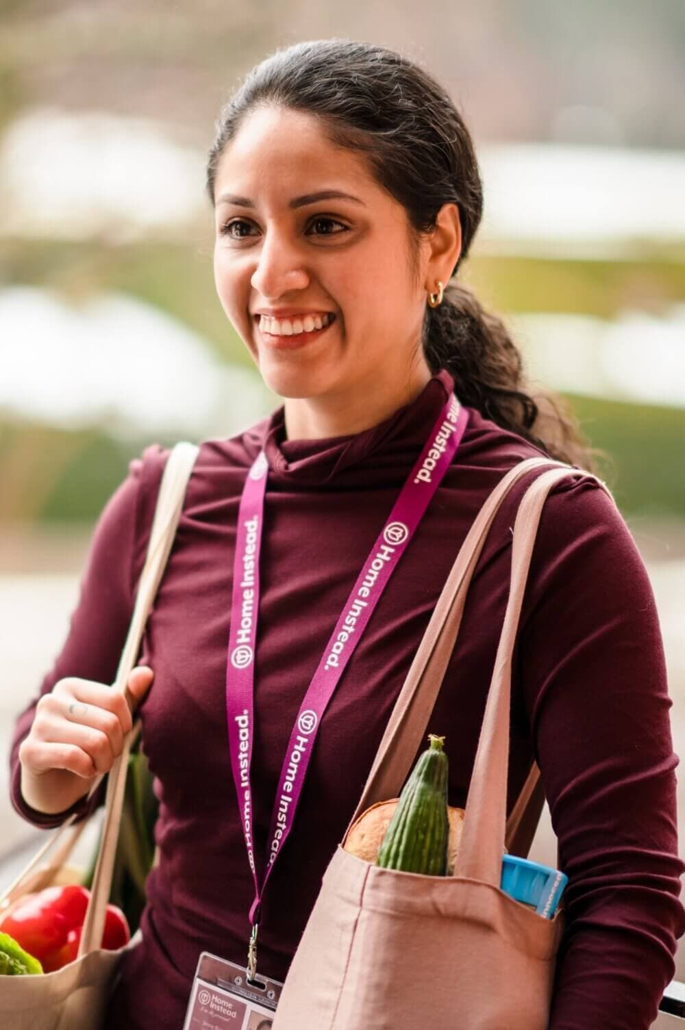 A smiling woman with a ponytail carries groceries in reusable bags, wearing a maroon shirt and a lanyard. - Home Instead