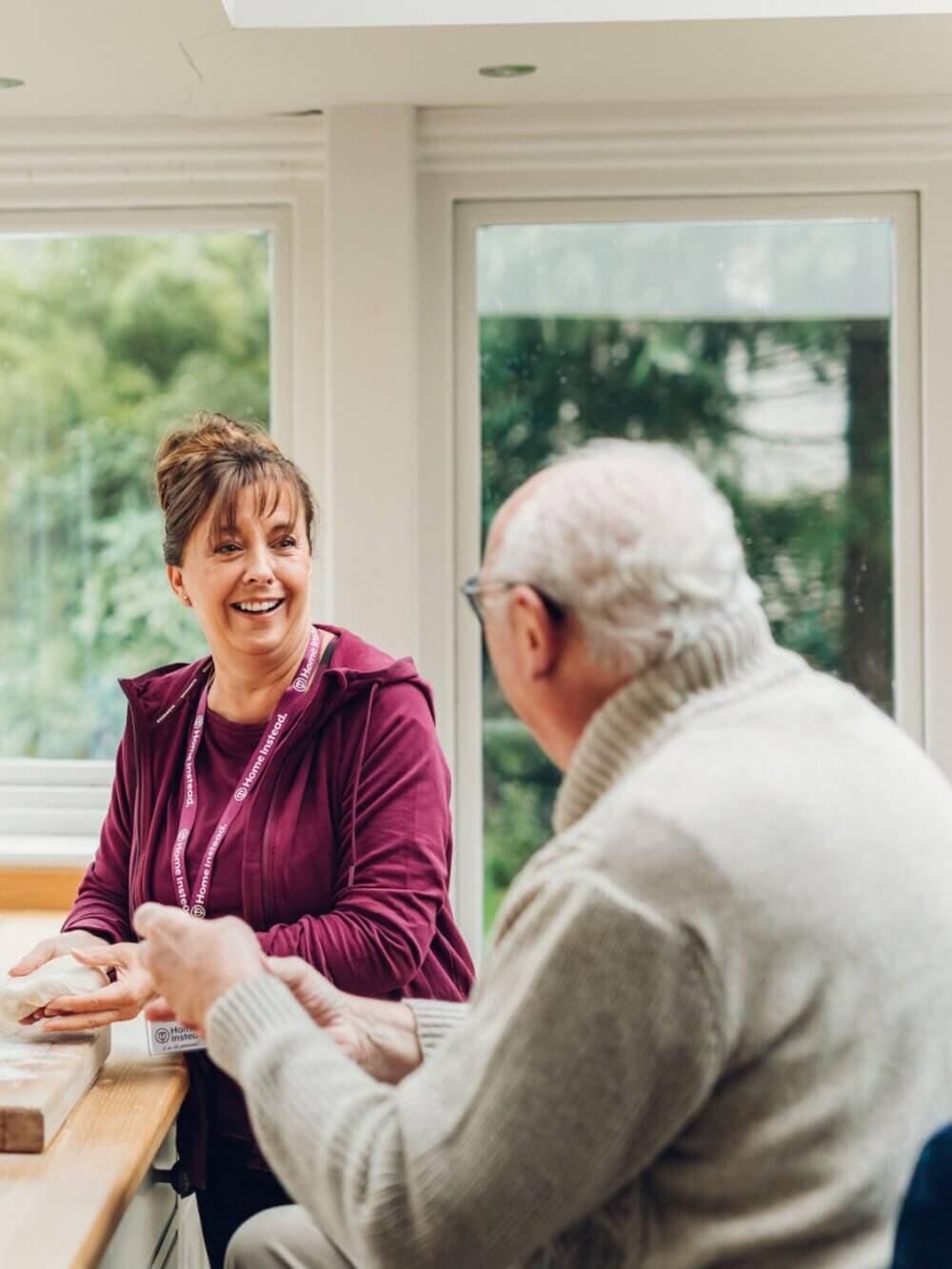 A caregiver smiles while talking to an elderly man sitting at a table in a bright room with large windows. - Home Instead