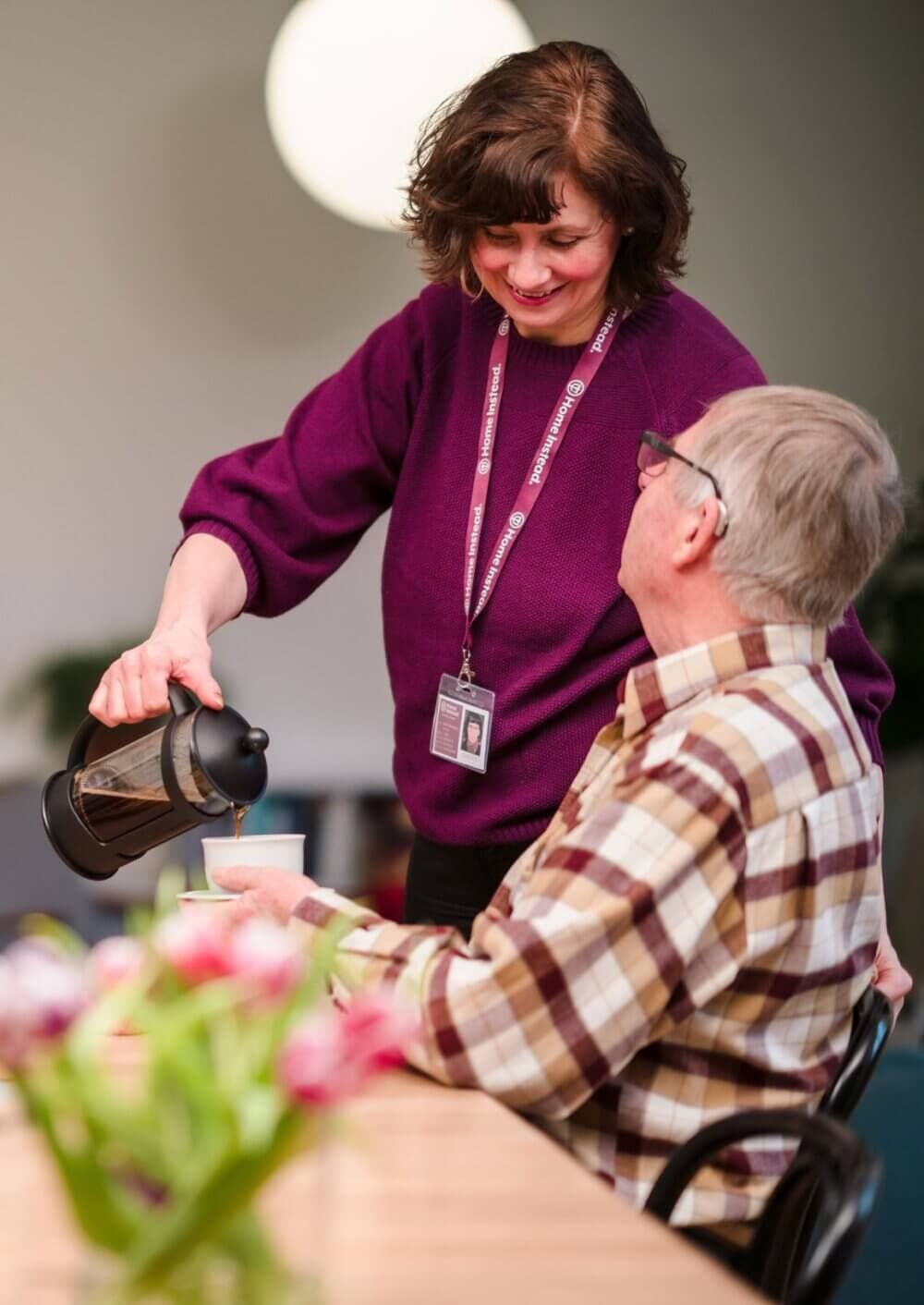 A woman wearing a lanyard pours coffee for an elderly man at a table with flowers. - Home Instead