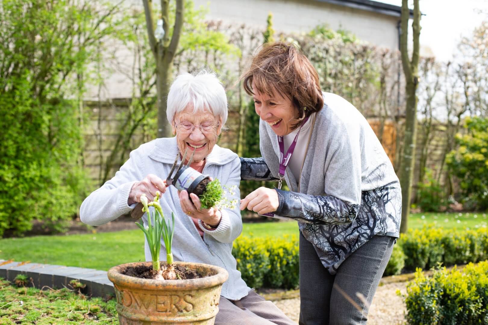 An elderly woman and a younger woman, both smiling, are planting flowers in a pot in a garden. - Home Instead