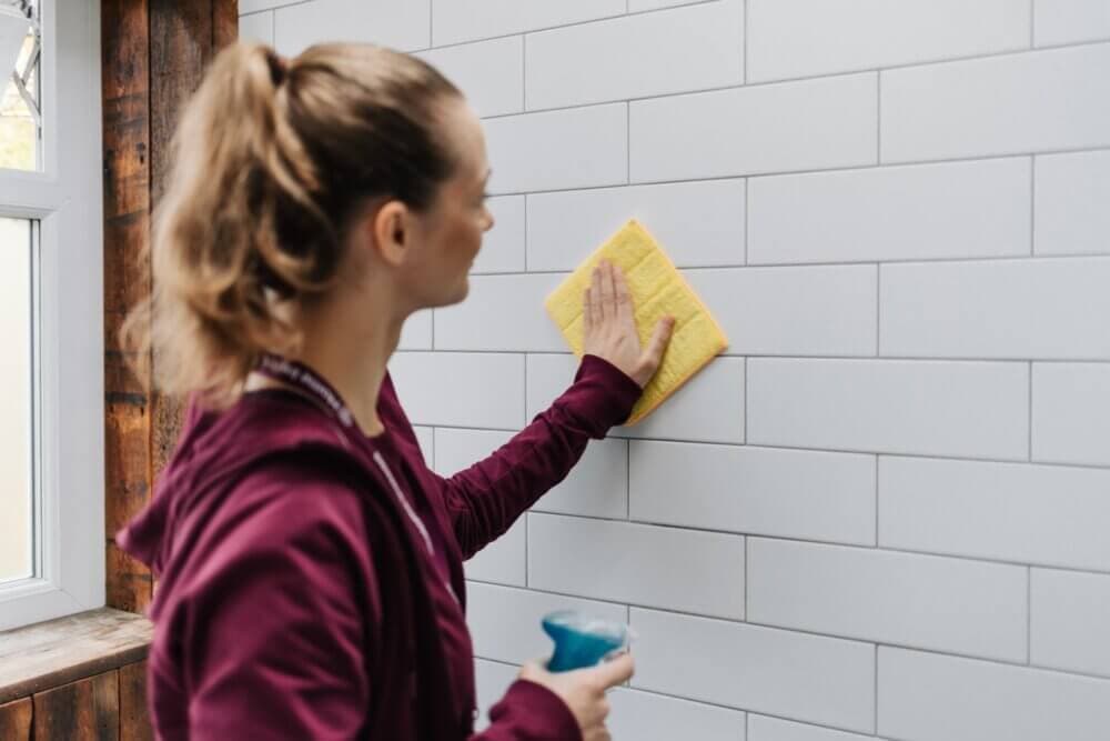 A person with a ponytail, in a red hoodie, cleans white tiled wall with a yellow sponge and blue spray bottle. - Home Instead