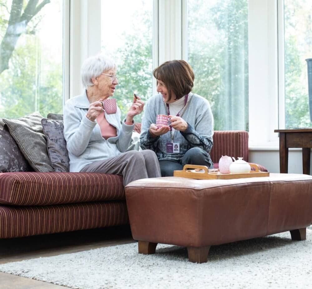 An elderly woman and a caregiver sit on a sofa, smiling and chatting while holding mugs in a bright, cosy room. - Home Instead Southampton