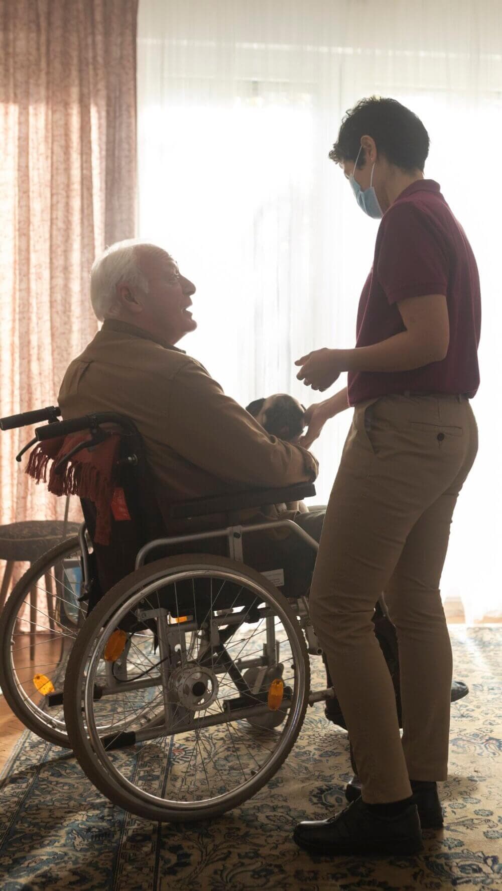 An elderly man in a wheelchair gets assistance from a healthcare worker wearing a mask in a warmly lit room. - Home Instead