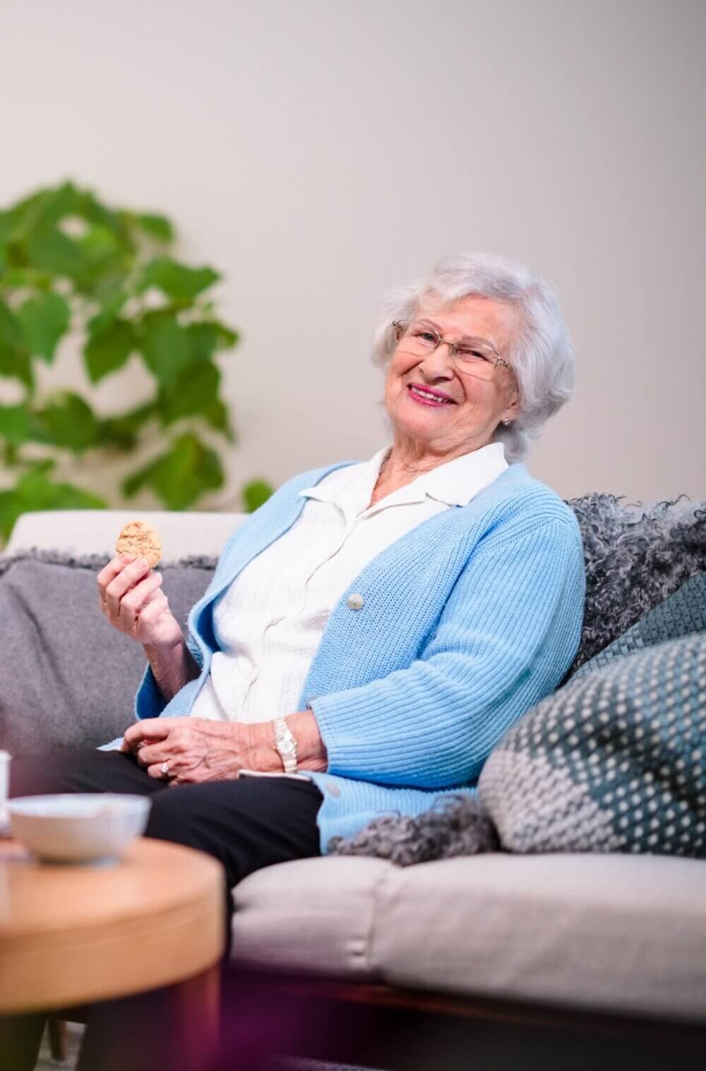 Elderly woman with white hair in a blue cardigan smiling and holding a cookie while sitting on a couch. - Home Instead
