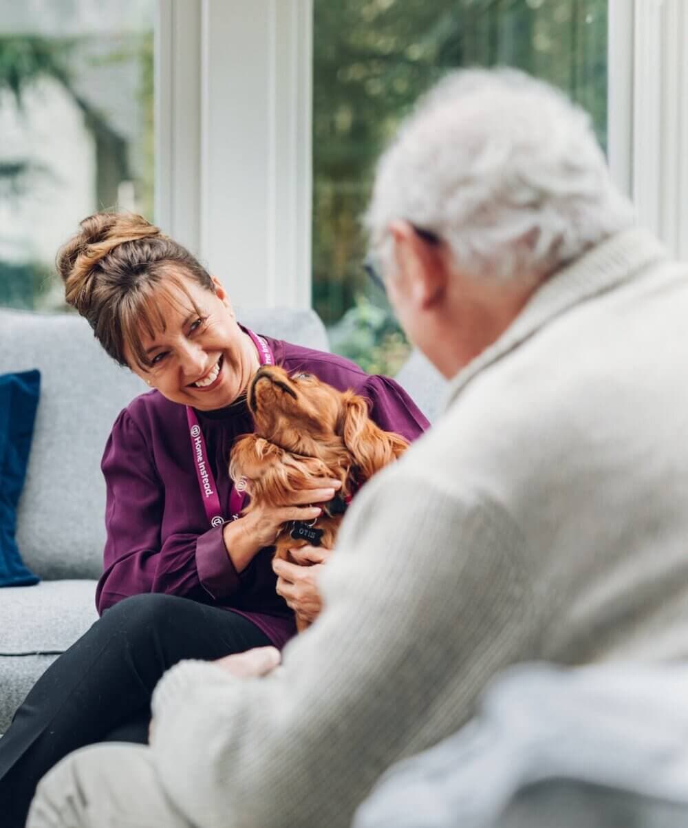 Person with a lanyard smiling, holding a dog, and sitting on a couch opposite an older man. - Home Instead