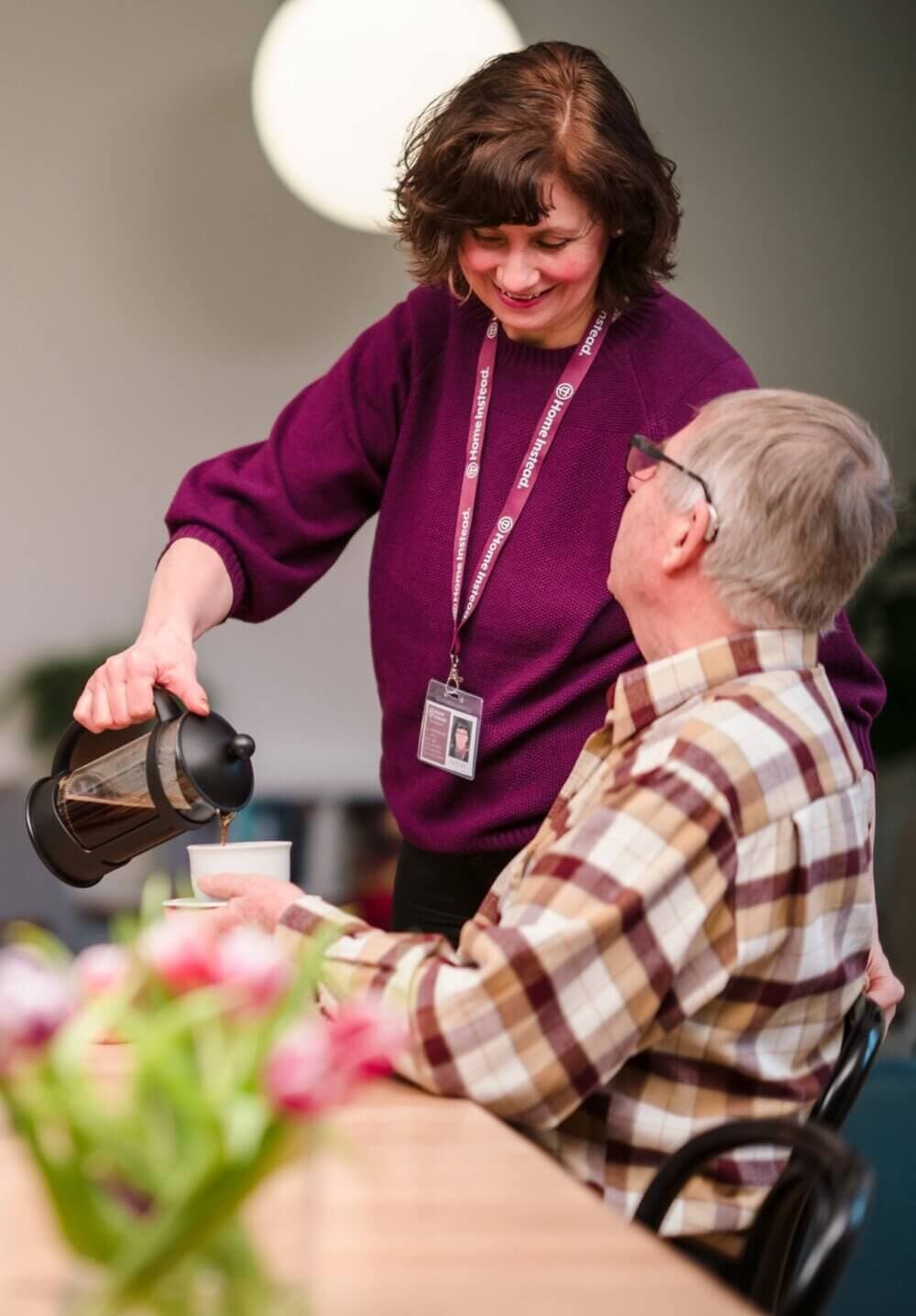 A woman in a purple sweater pours coffee for an older man wearing a plaid shirt at a table with a flower centerpiece. - Home Instead