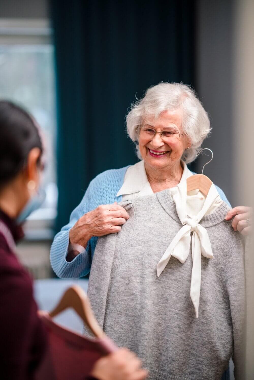 An elderly woman holds up a gray sweater with a white bow, smiling at another person who is holding a different sweater. - Home Instead