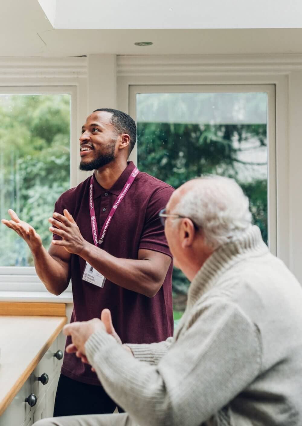 A smiling man chats with an elderly man in a cozy room with large windows and a view of greenery outside. - Home Instead