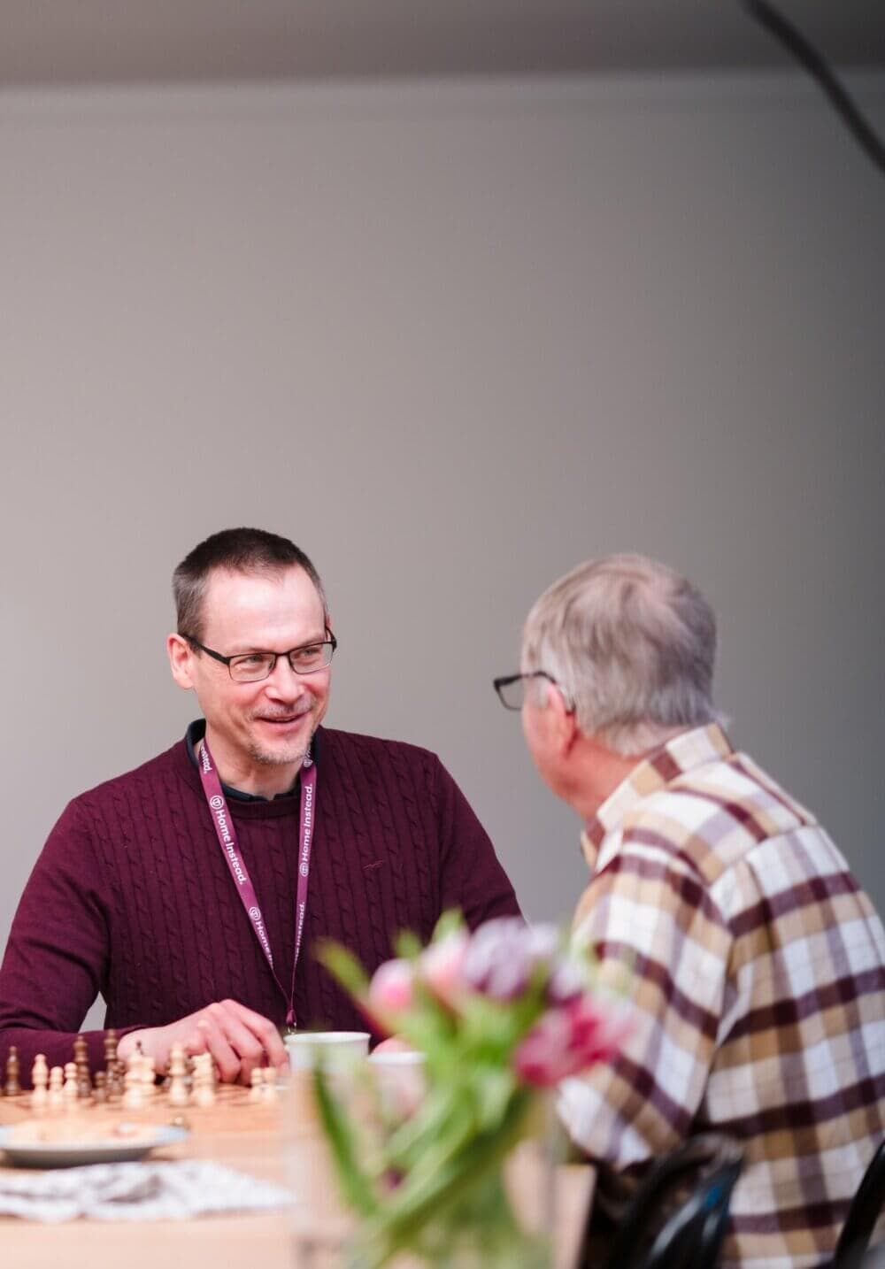 Two men are seated at a table, deep in conversation, with a chessboard and flowers in the foreground. - Home Instead