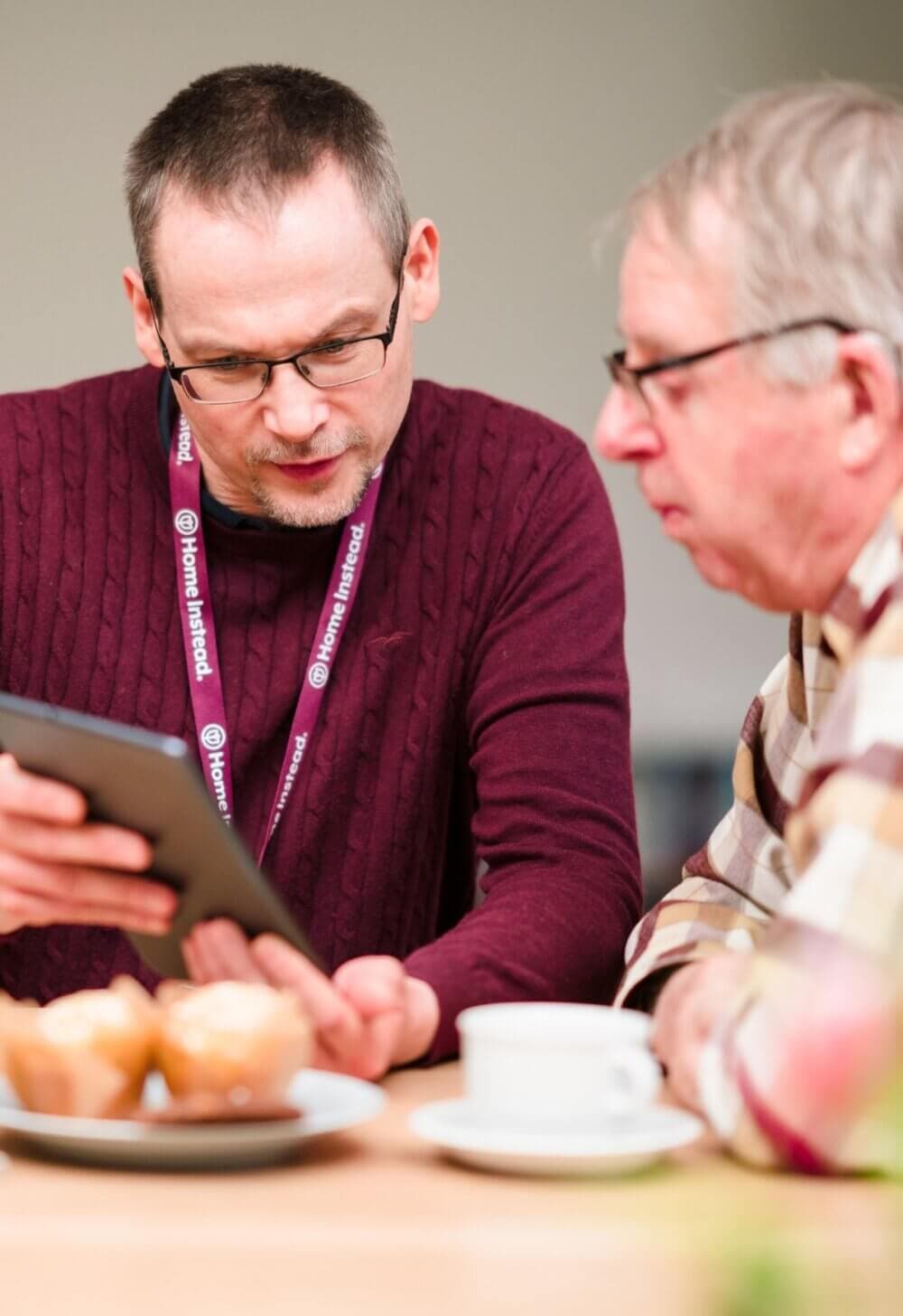 Two men sitting at a table with muffins, one showing a tablet to the other. Both wear glasses and name tags. - Home Instead Bournemouth & Christchurch