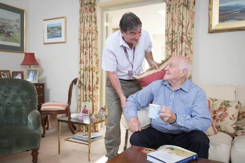 A man stands, smiling, over an elderly man seated with a mug on a couch in a comfortable, decorated living room. - Home Instead