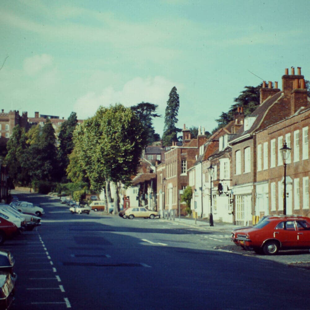 A quiet street lined with parked cars and red brick buildings, with trees and a large structure in the background. - Home Instead