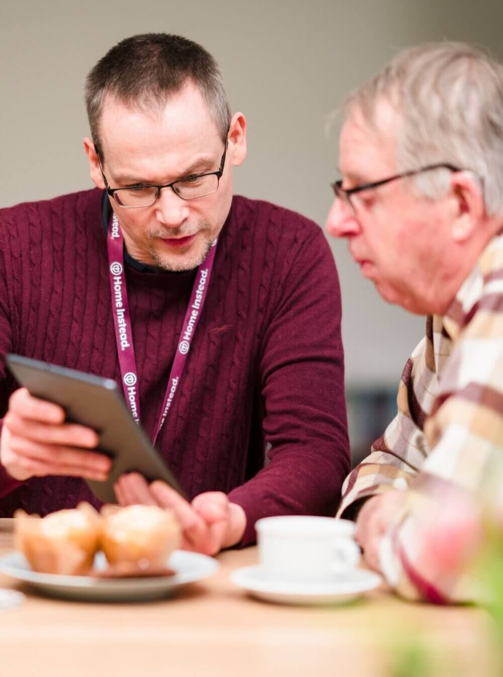 Two men sit at a table with muffins and a cup. One man shows the other something on a tablet. - Home Instead