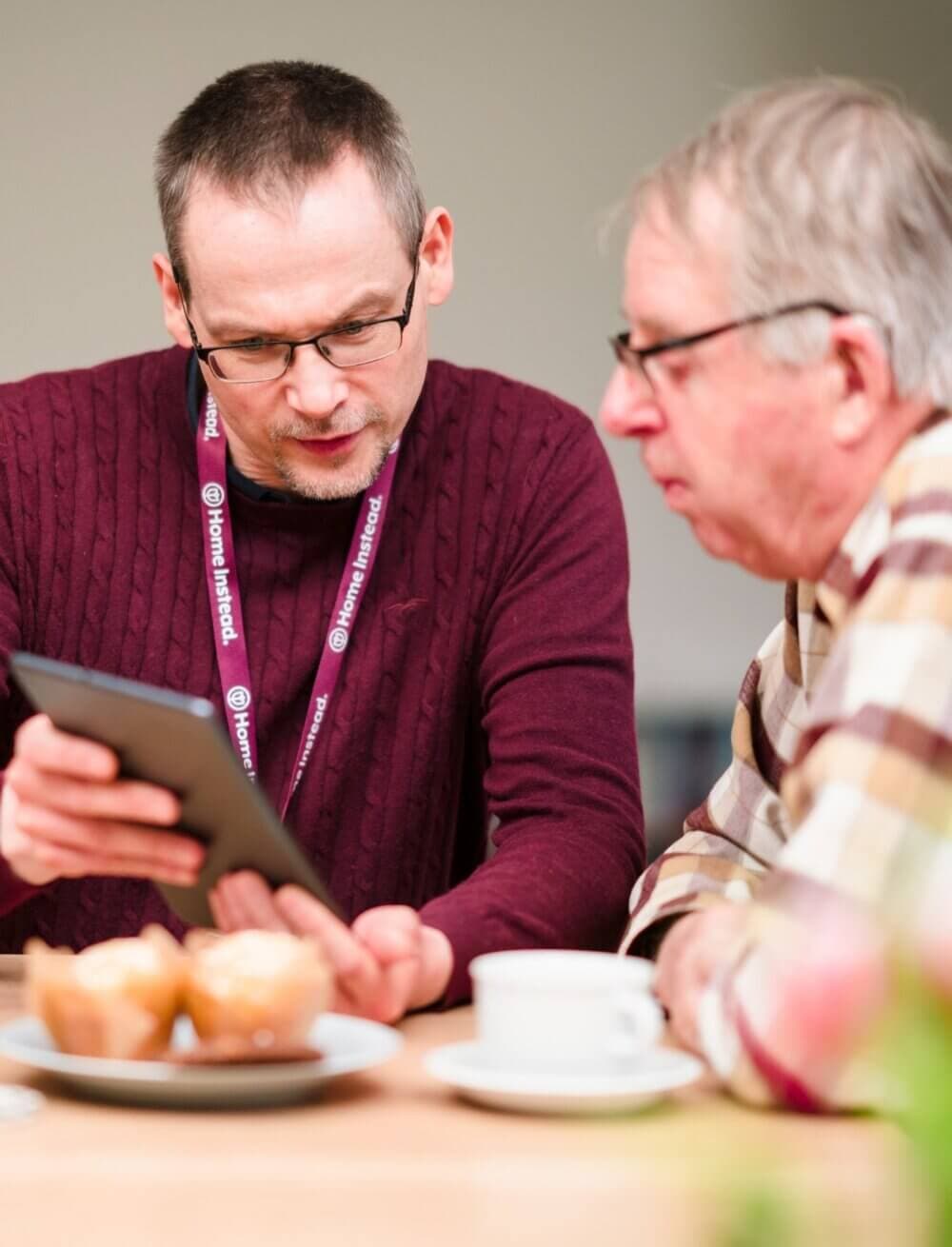 Two men sit at a table, one holding a tablet, with coffee and muffins in the foreground. - Home Instead