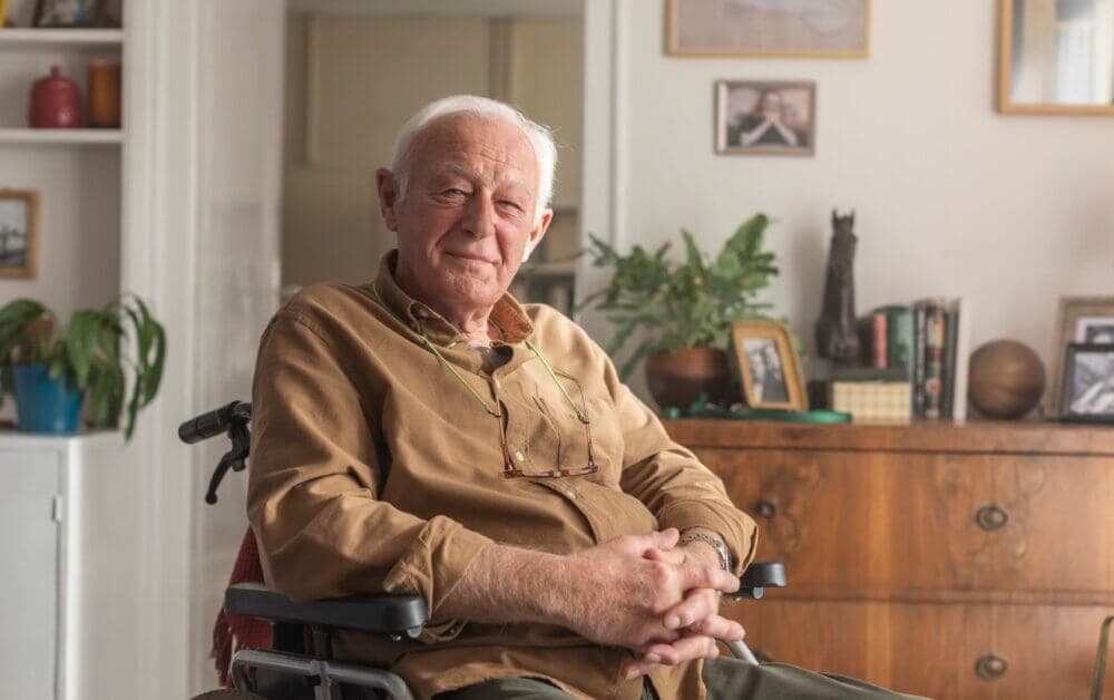 An elderly man sitting in a wheelchair, smiling in a cozy room with plants, pictures, and a wooden dresser in the background. - Home Instead