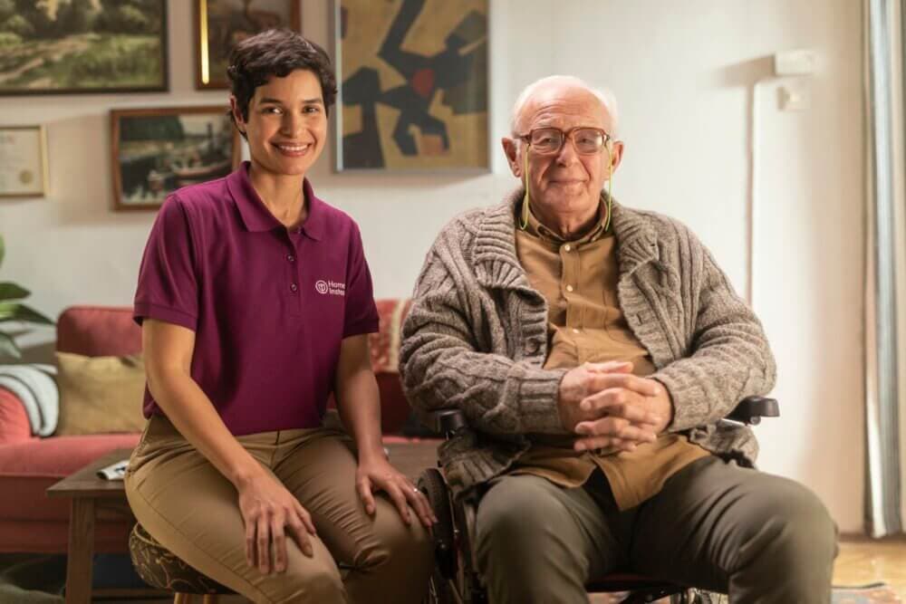Smiling caregiver in a purple shirt sits beside an elderly man in a wheelchair in a cozy, art-filled living room. - Home Instead