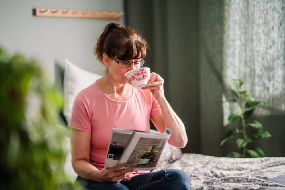 Woman in a pink shirt, wearing glasses, drinks from a cup while reading a magazine in a cozy room with plants. - Home Instead