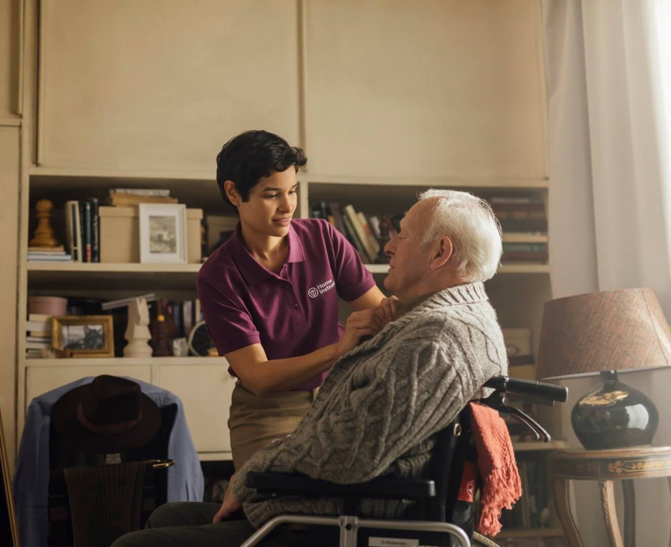 A caregiver in a purple shirt assists an elderly man in a wheelchair in a cozy room with bookshelves and a lamp. - Home Instead