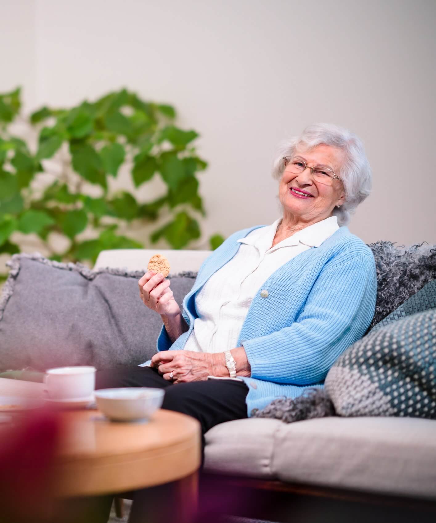 Elderly woman with white hair, in a blue cardigan, smiling and holding a cookie while sitting on a couch. - Home Instead