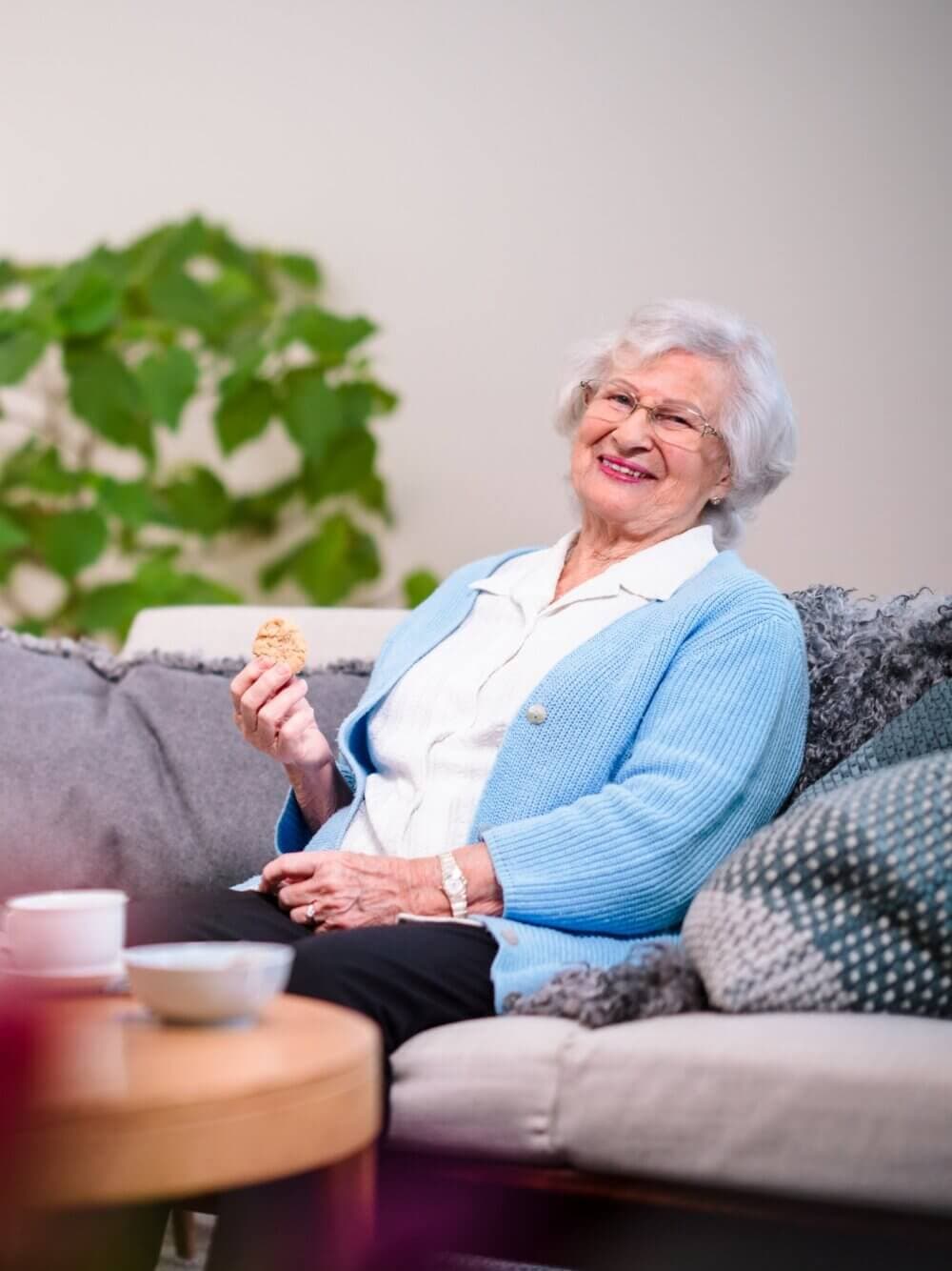Elderly woman sitting on a couch, smiling, and holding a cookie. A cup and bowl are on the table in front of her. - Home Instead