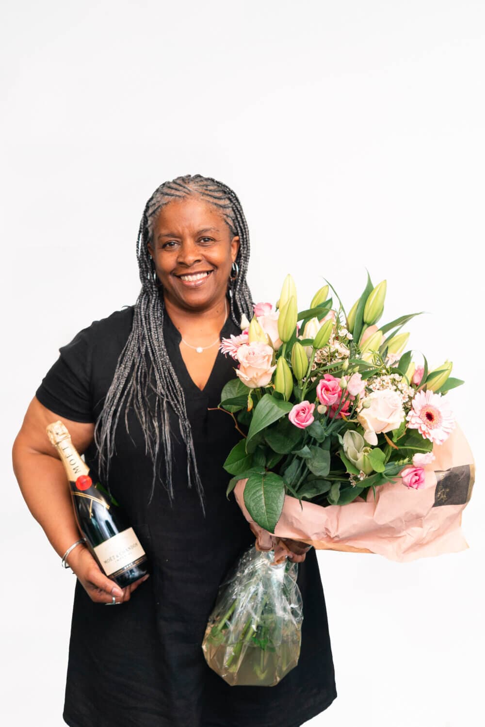 Woman smiling, holding a bouquet and a bottle of sparkling wine against a plain white background. - Home Instead