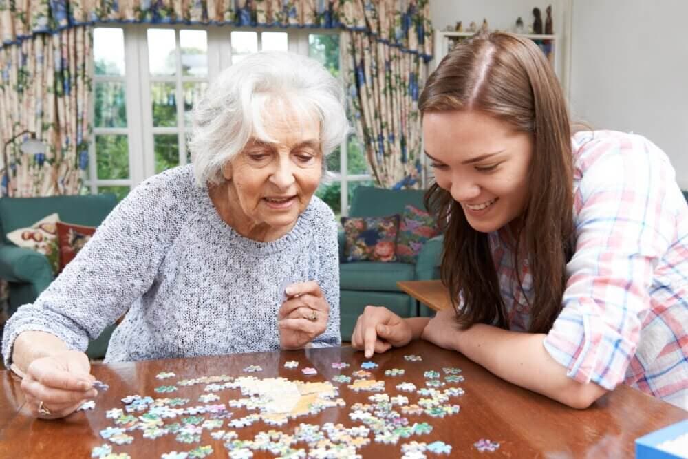 An elderly woman and a young woman are smiling and working together on a jigsaw puzzle at a wooden table. - Home Instead